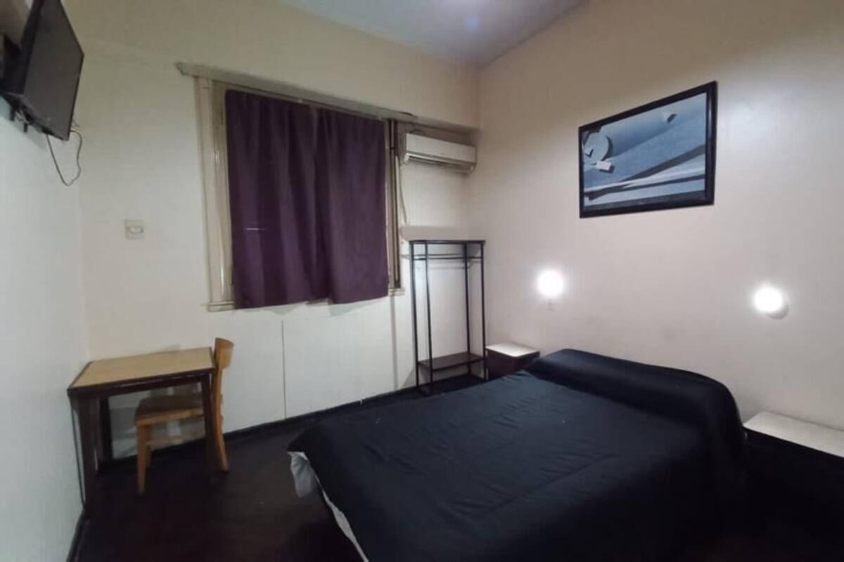 A small, simple room with a bed, a desk, a chair, and a window with curtains.