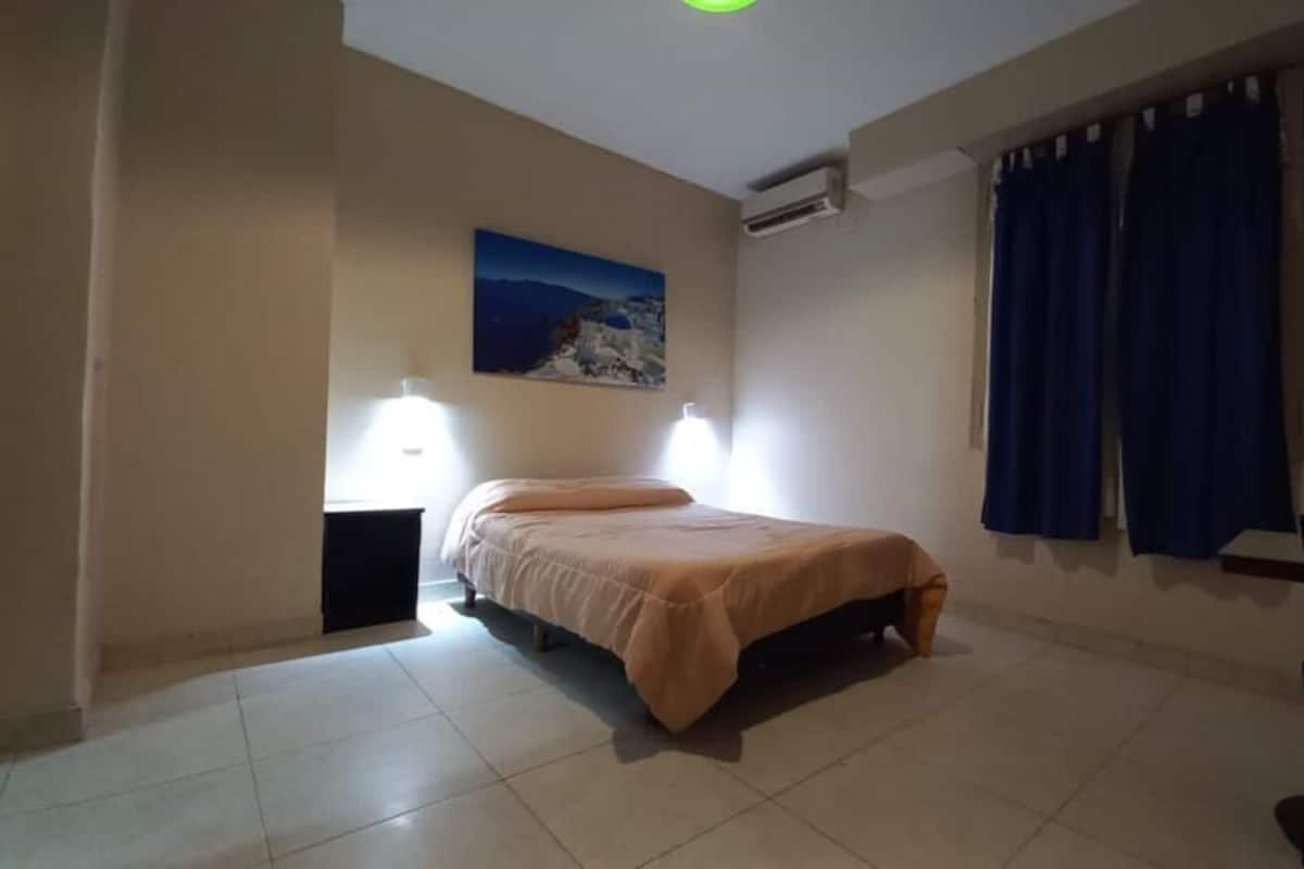 A bedroom with a bed, a nightstand, a chair, a painting on the wall, and a window with curtains.