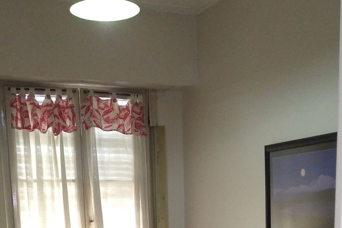 A room with two single beds, a window with sheer curtains, a wall-mounted picture, and a hanging light fixture.
