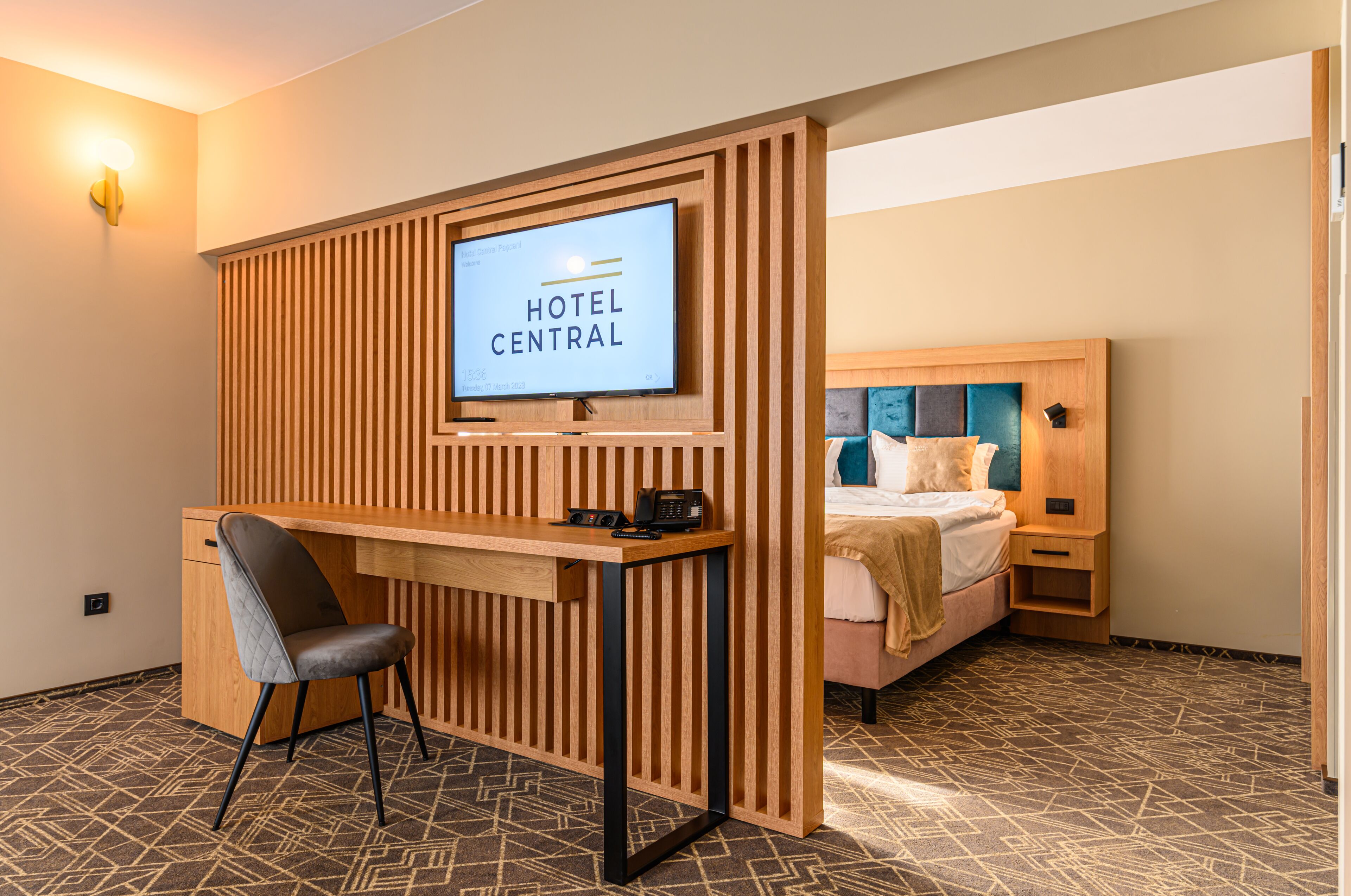 Una habitación de hotel con un escritorio de madera, una cama con cabecera azul y un televisor que muestra “HOTEL CENTRAL”.