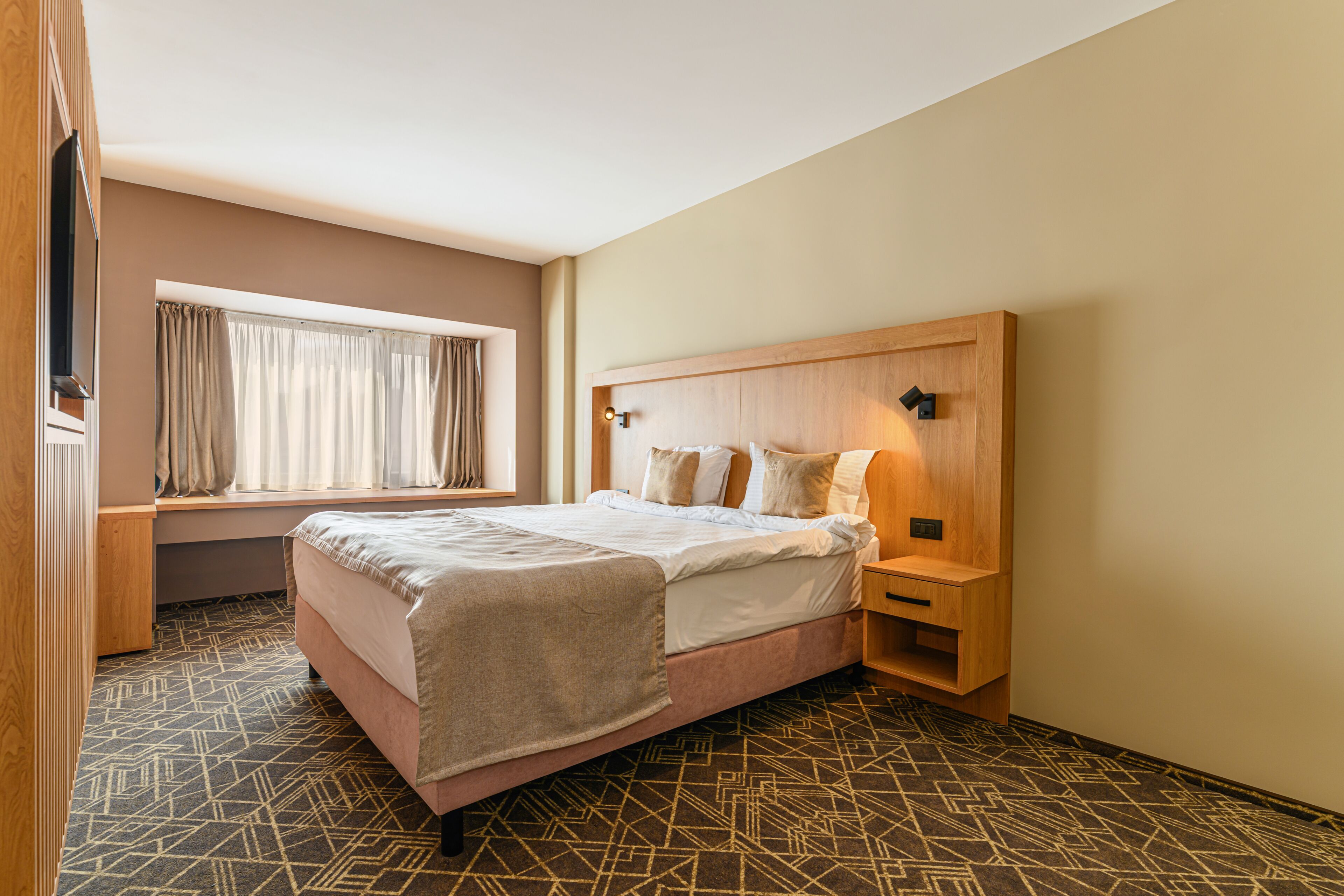 Habitación de hotel ordenada con cama, mesita de noche, cortinas y alfombra estampada.