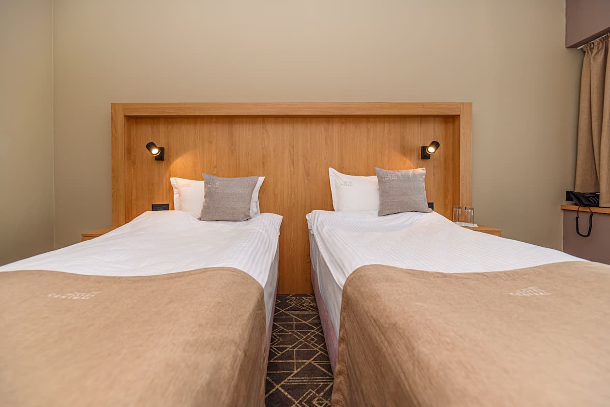 A hotel room with two single beds, a wooden headboard, bedside lamps, and a small nightstand with a lamp.