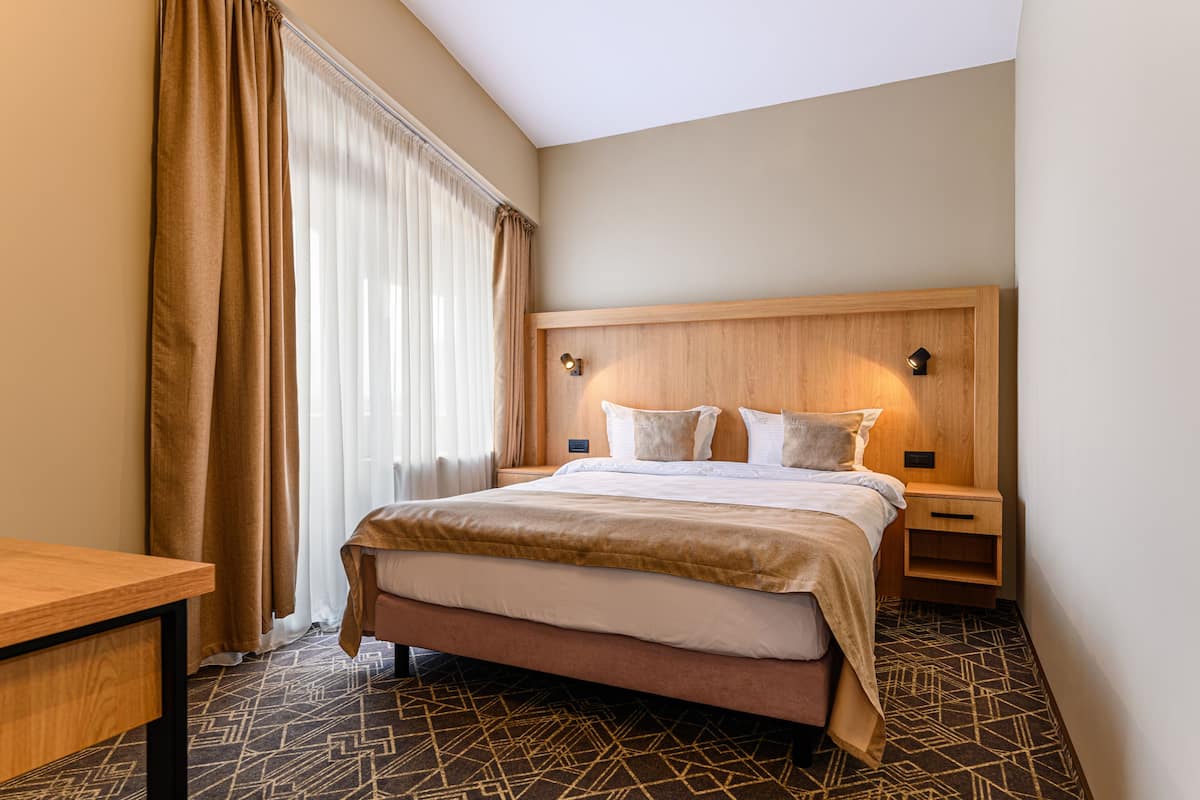 A hotel room with a bed, a nightstand, a wooden headboard, a desk, and a patterned carpet.