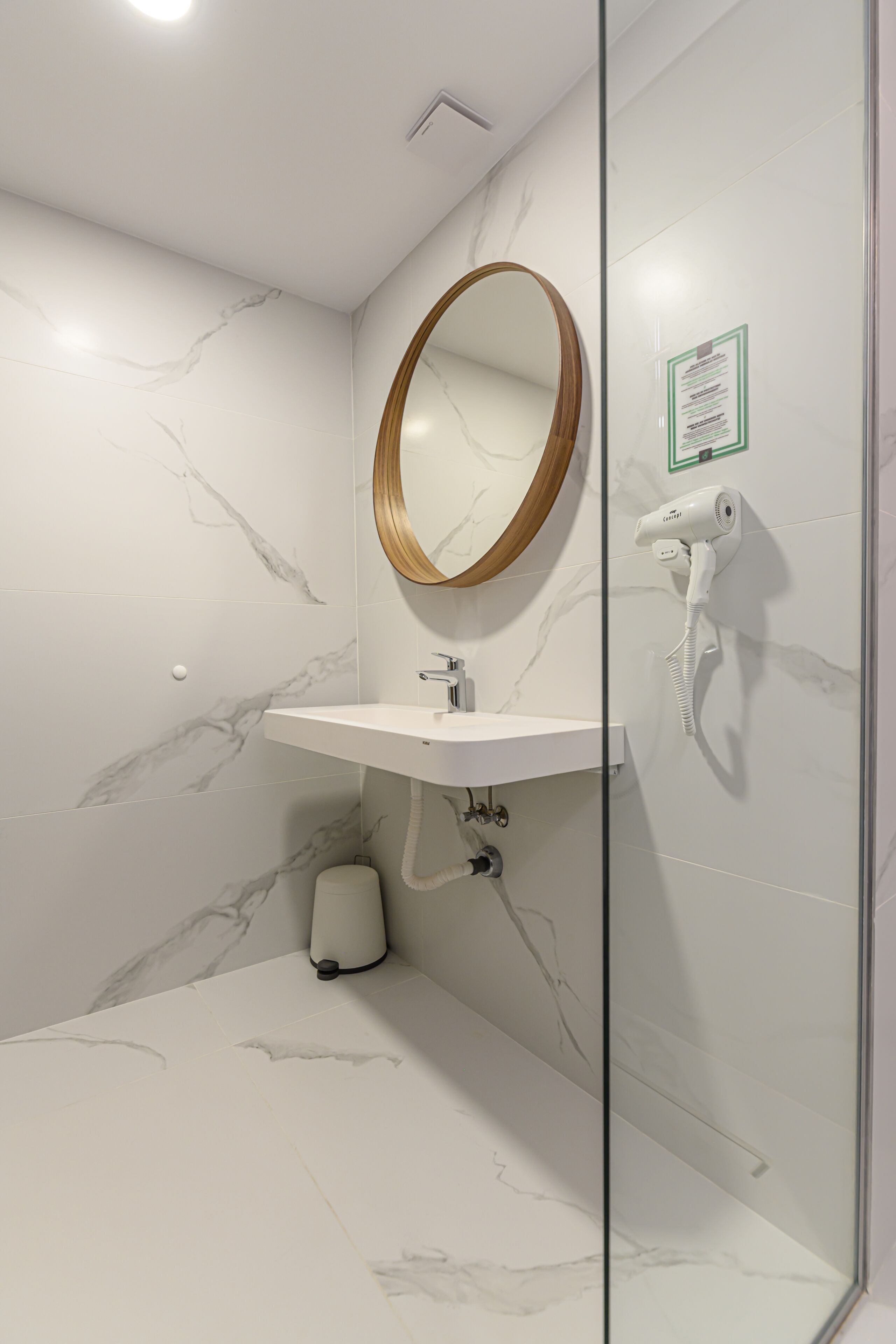 Un baño moderno con pared de mármol, un lavabo blanco, un espejo con marco de madera y un secador de pelo montado en la pared.