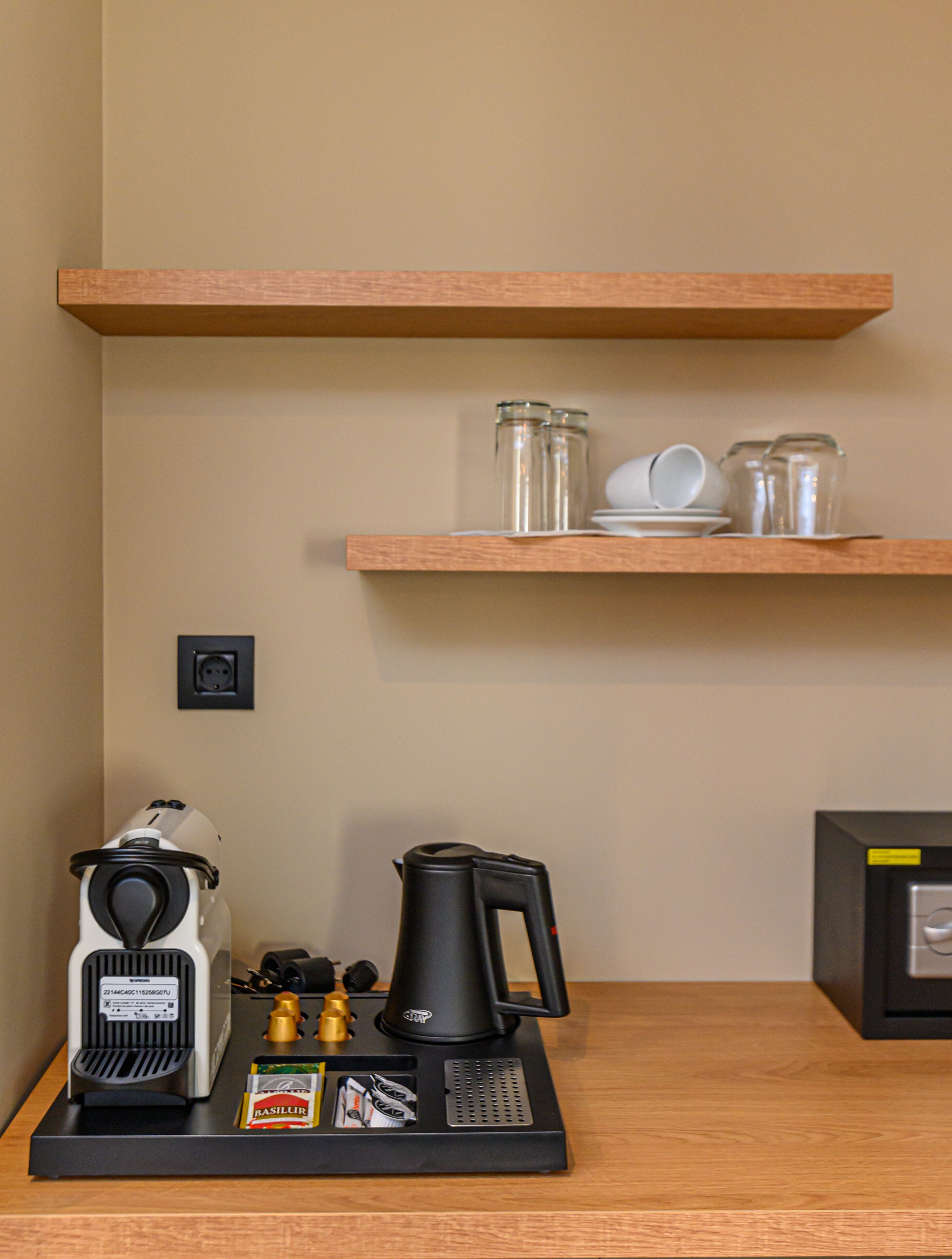 A kitchenette with a coffee maker, a kettle, and salt and pepper shakers on a black tray. Above the tray, there are two wooden shelves holding glass jars and white cups.
