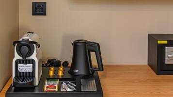 A kitchenette with a coffee maker, a kettle, and salt and pepper shakers on a black tray. Above the tray, there are two wooden shelves holding glass jars and white cups.