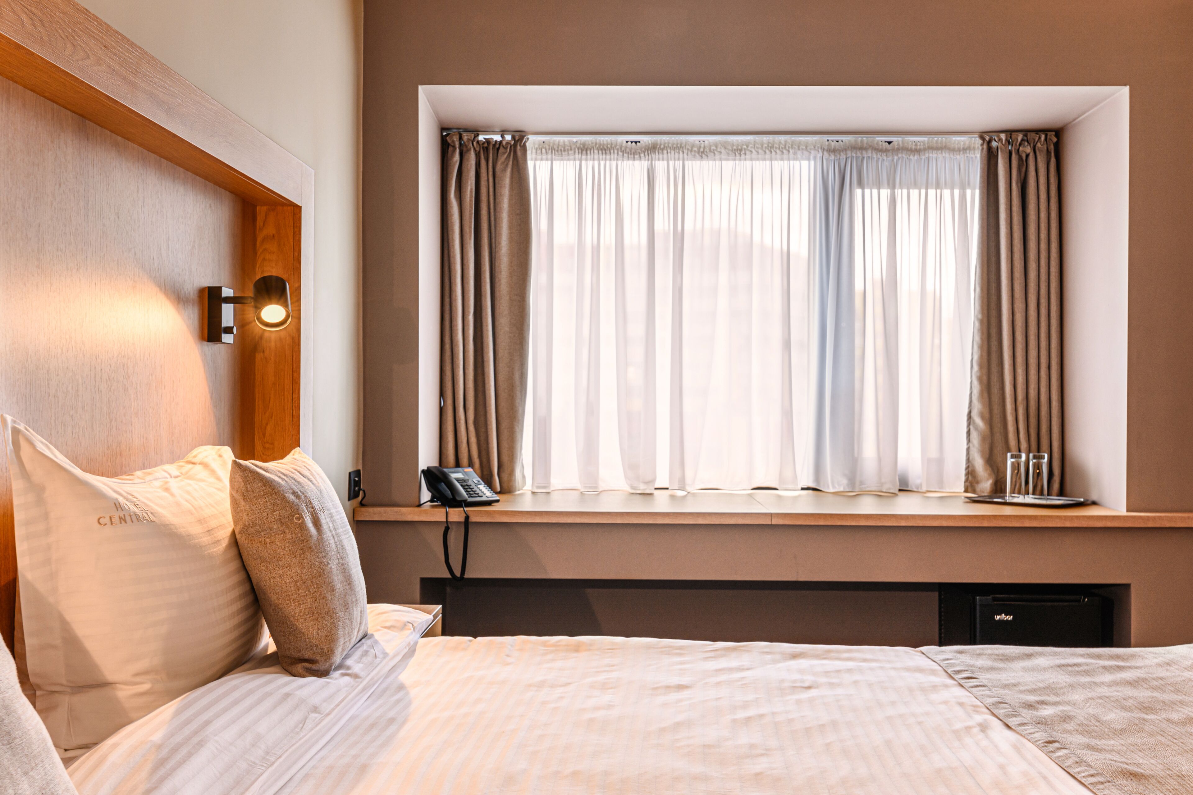 Habitación de hotel con cama, teléfono en la mesita de noche, ventana con cortinas y lámpara fijada a la pared.
