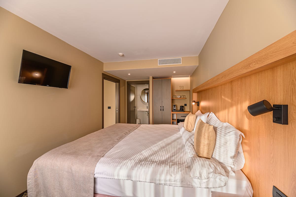 A hotel room with a large bed, a flat-screen TV mounted on the wall, a wooden headboard, and a small shelf with items.