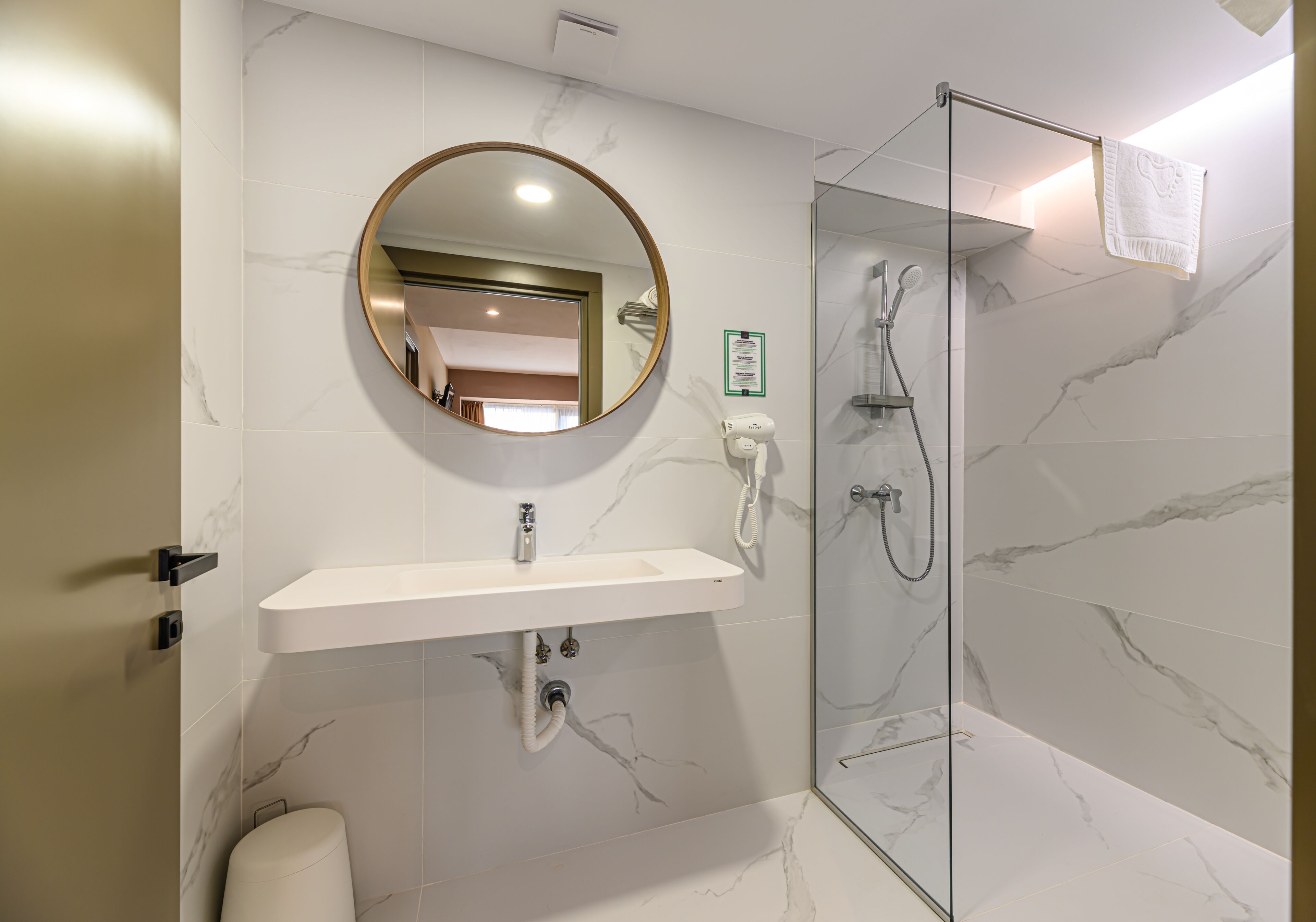 Un baño moderno con pared de mármol, espejo redondo, lavamanos blanco y cabina de ducha con mampara de vidrio.