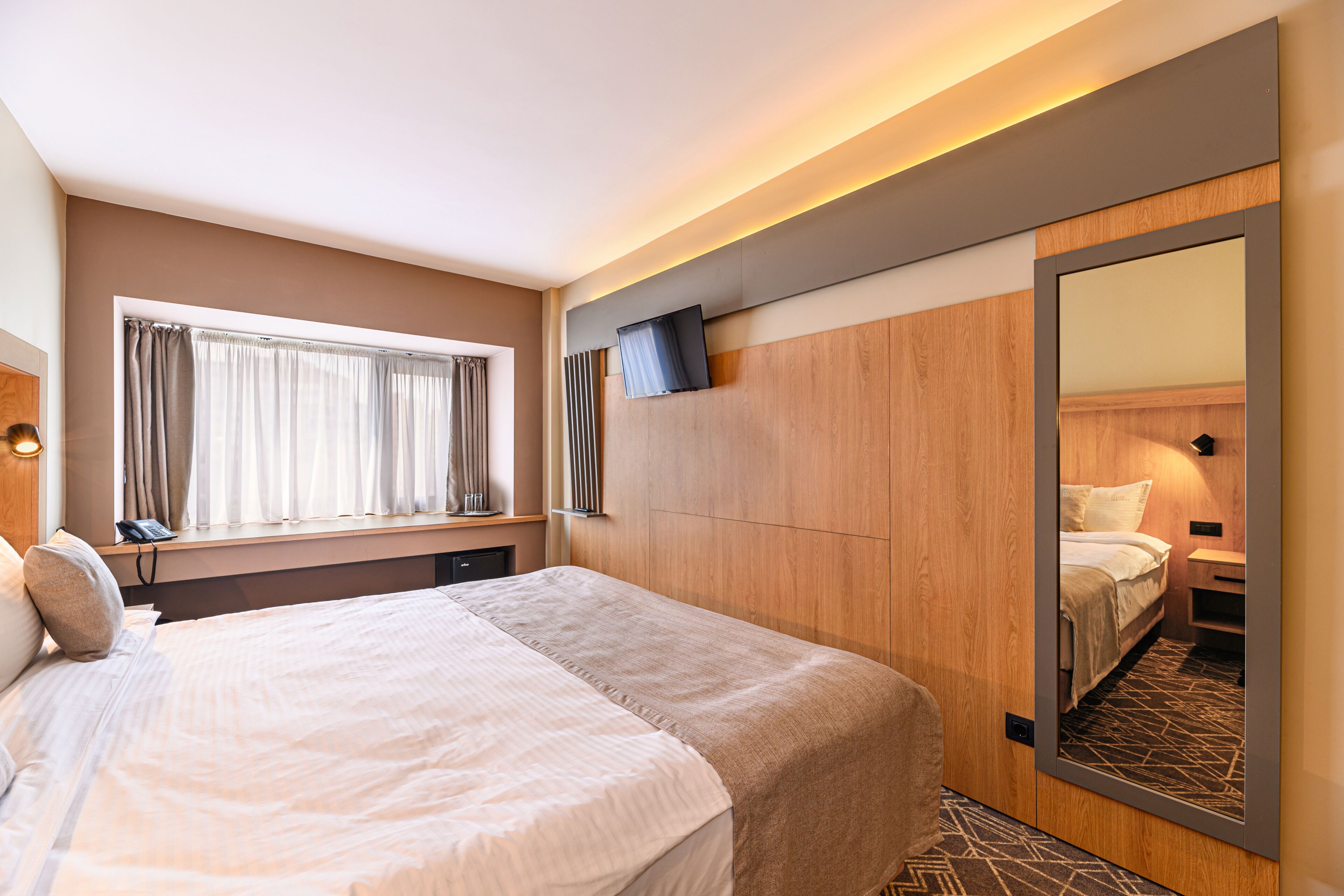 A modern hotel room with a large bed, a bedside table, a mirror, and a window with curtains.