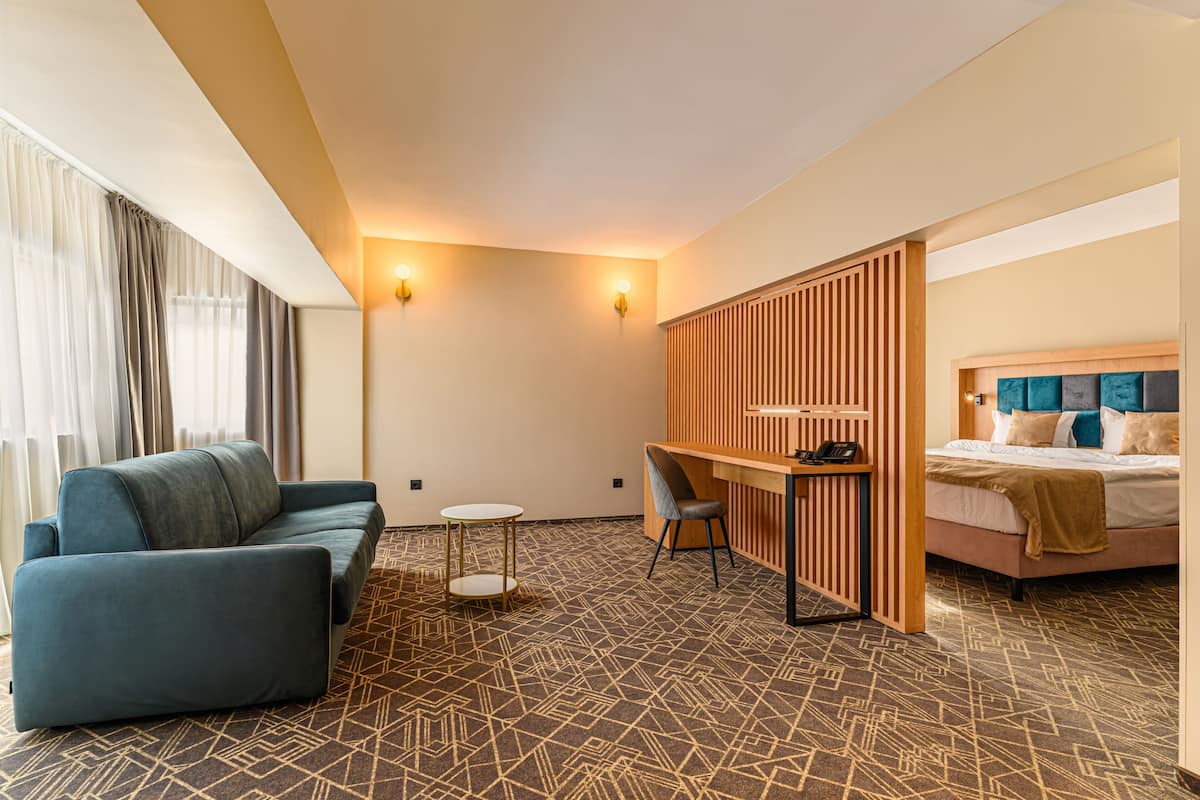 A hotel room with a blue sofa, a small round table, a desk with a chair, a bed with a headboard, and a patterned carpet.