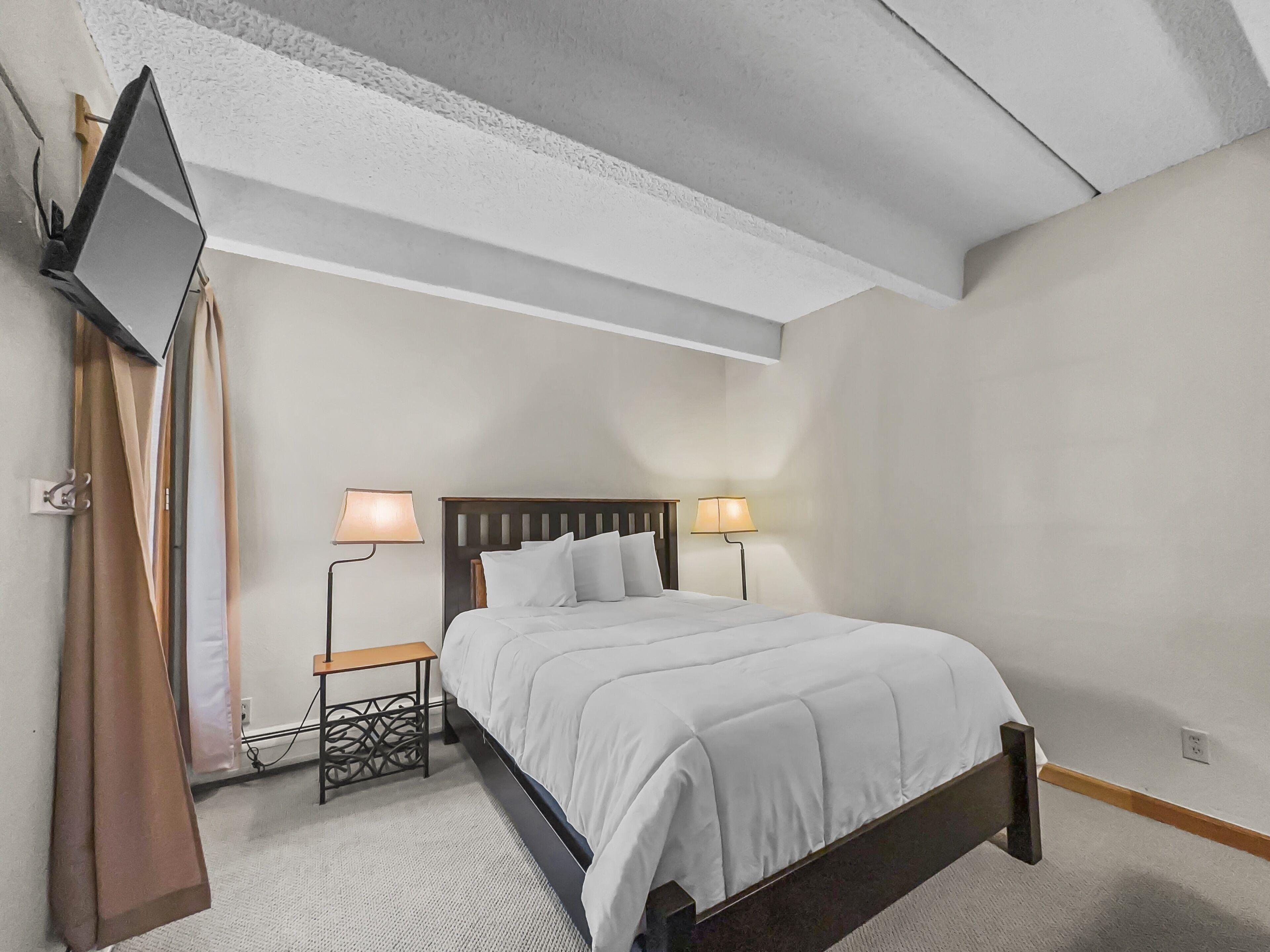Une chambre d’hôtel bien aménagée, avec un lit, deux tables de chevet, un téléviseur à écran plat fixé au mur et un plafond avec des poutres apparentes.