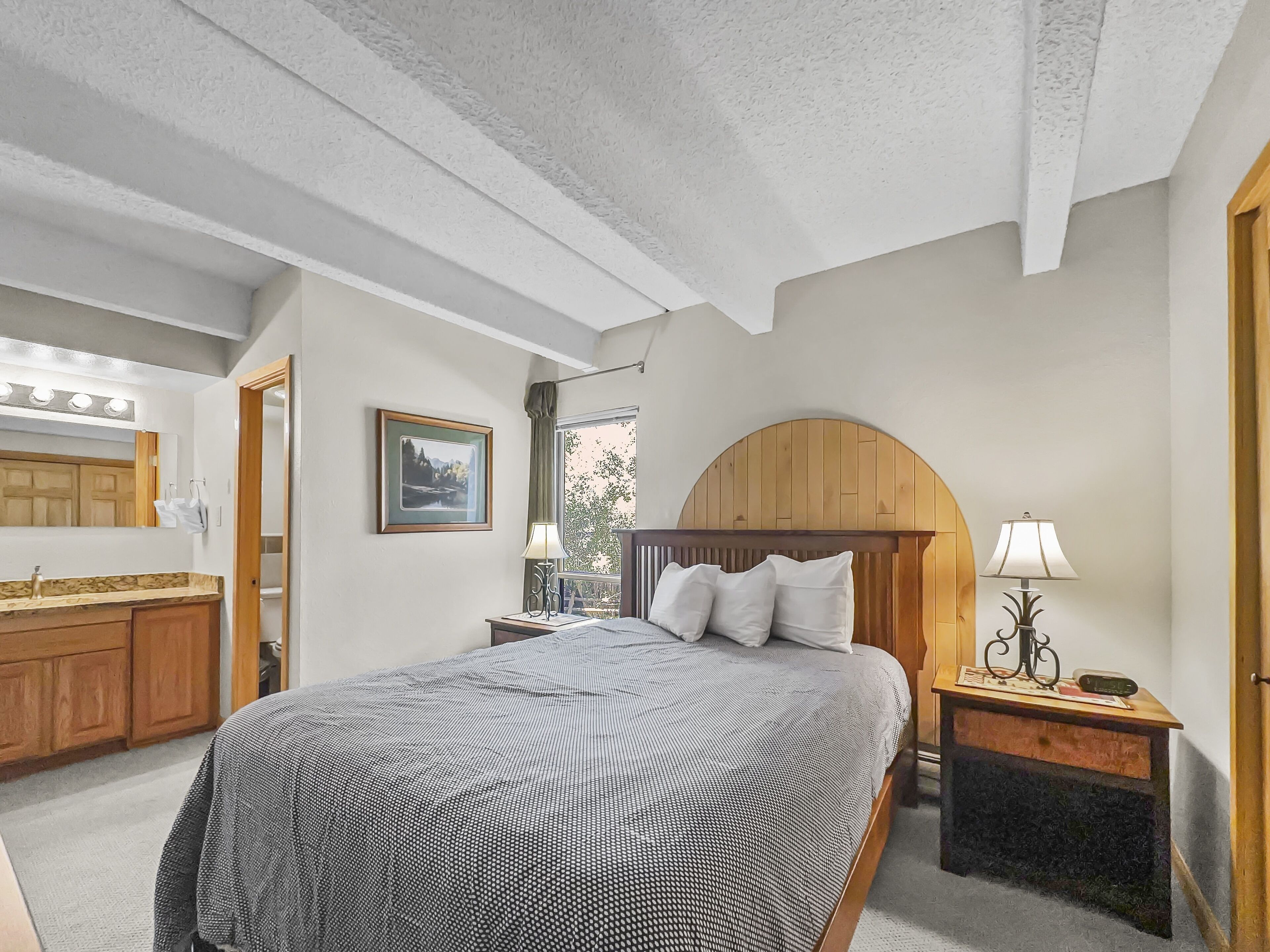 Ein Schlafzimmer mit einem großen Bett, einem Holz-Kopfteil, Nachttischen, einer Nachttischlampe, einem gerahmten Bild an der Wand und einem Badezimmer mit Spiegel und Schminktisch.