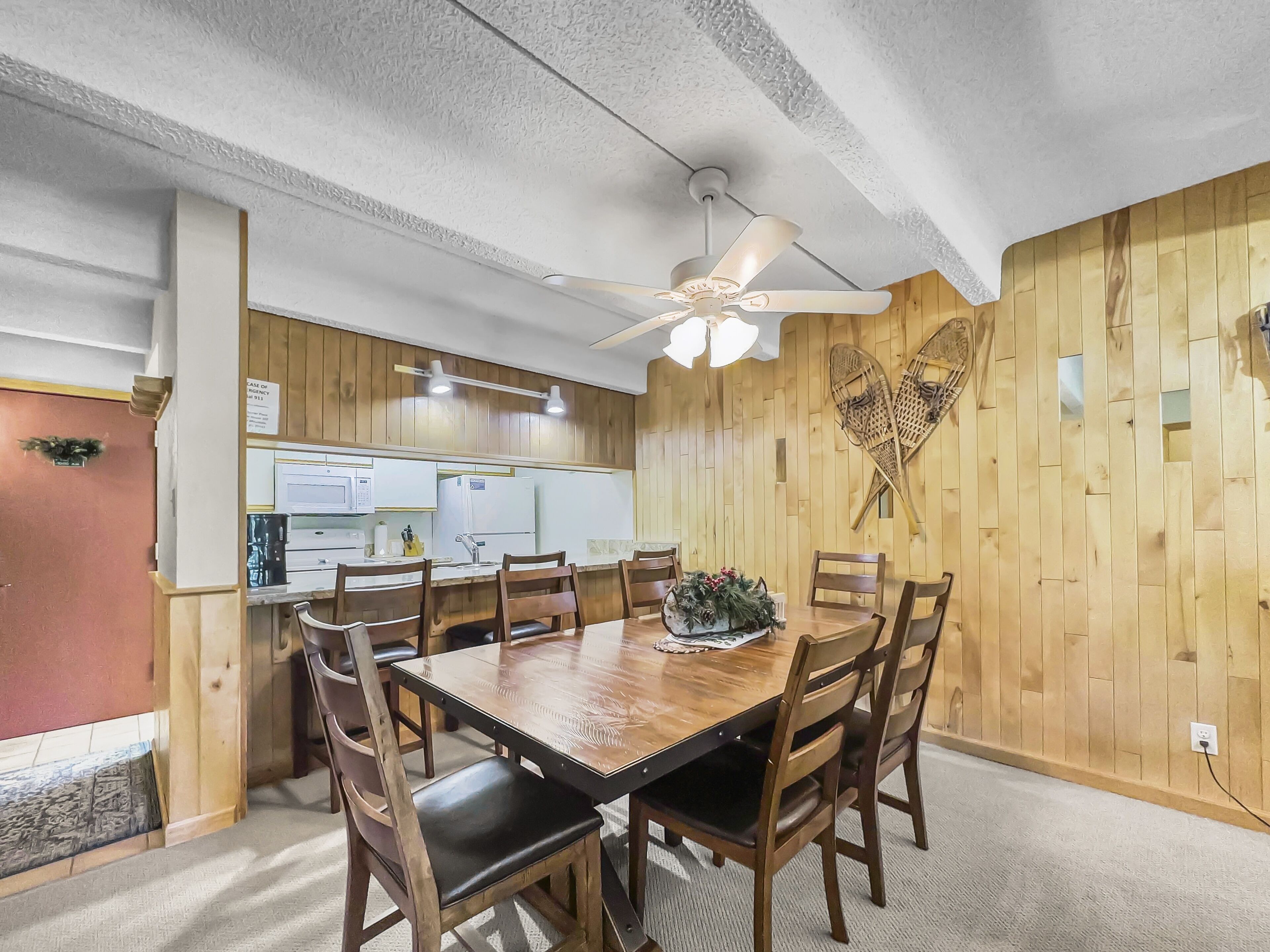 A dining area with a wooden table and chairs, a ceiling fan, and a kitchen in the background.