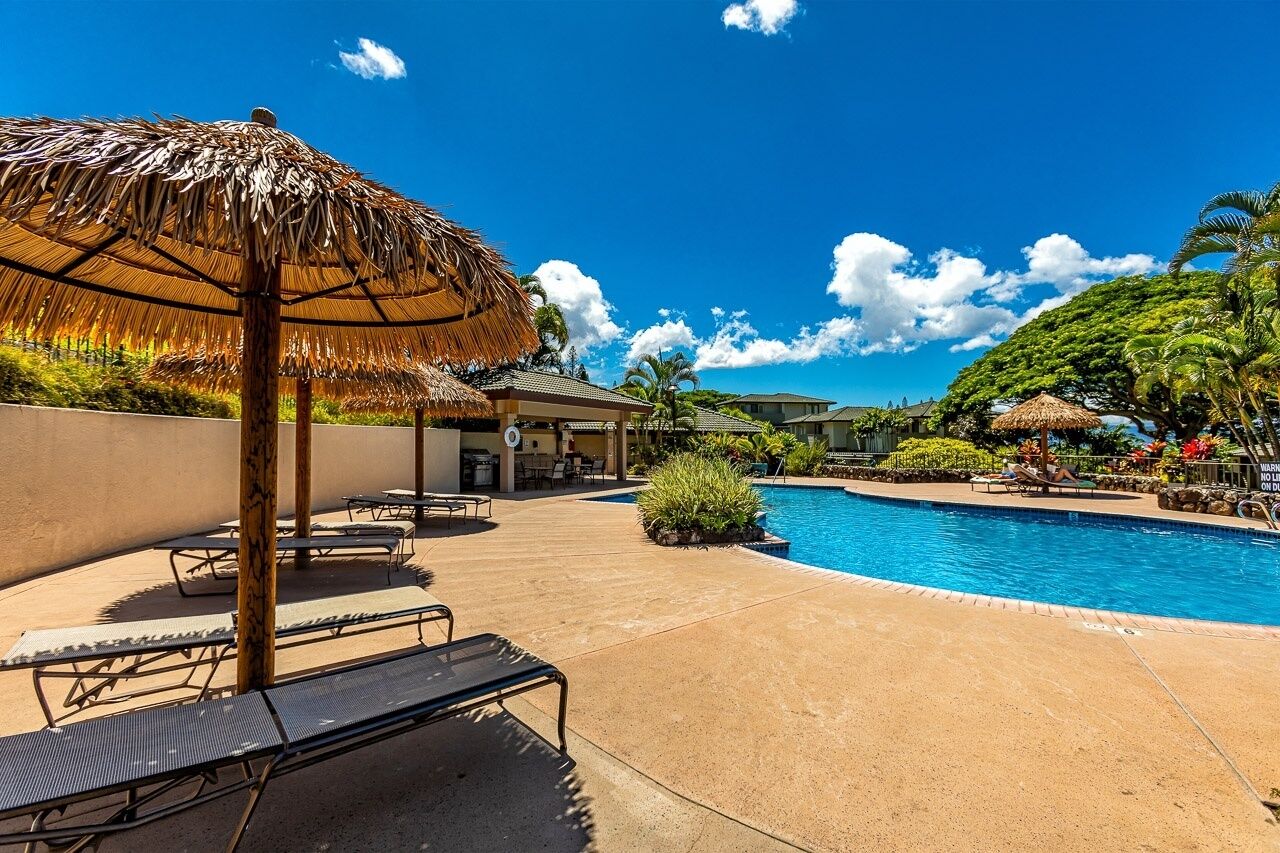 A pool area with lounge chairs and a thatched umbrella, surrounded by greenery and a clear blue sky.