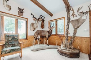 Una habitación con paredes de madera, un ciervo disecado, una silla con tapicería alusiva a la naturaleza y una ventana con vistas a árboles.