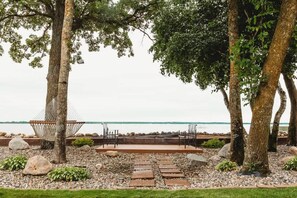 Un espacio al aire libre tranquilo con una hamaca, sillas y un hogar para fogata, rodeado de árboles y un sendero de piedras.