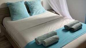 A neatly made bed with blue pillows, a white bedspread, and two rolled towels on a blue bed runner.