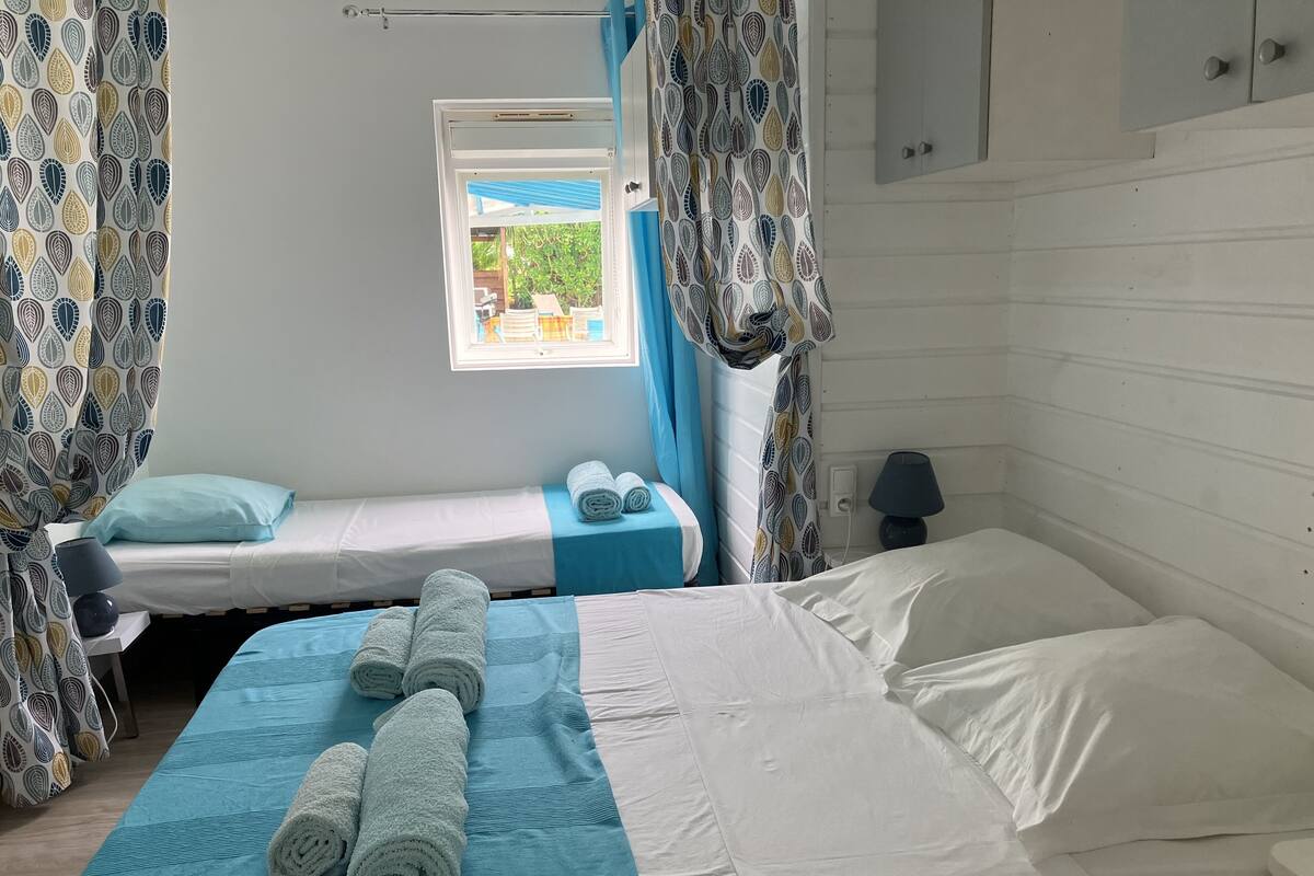 Une chambre avec deux lits, l’un recouvert d’une couverture bleue et l’autre d’une couverture blanche. Il y a des serviettes sur les lits et une fenêtre avec des rideaux.