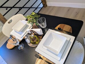 A dining table set with white plates, cutlery, and glassware, accompanied by a bottle of olive oil, a glass, and a potted plant.