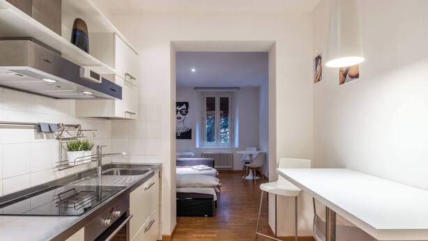 A compact kitchen with a stove, sink, and white cabinets. A small dining area with a table and chairs is visible in the background. - Maison Chanel (Bologna)