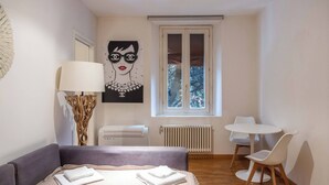 A bedroom with a bed, a bedside table, a lamp, a radiator, a window, and a small dining area with chairs. - Maison Chanel (Bologna)