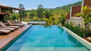 A rectangular pool with clear water, surrounded by wooden deck and lounge chairs, set against a backdrop of lush greenery and a clear sky. - Valle Paradiso (Imbituba)