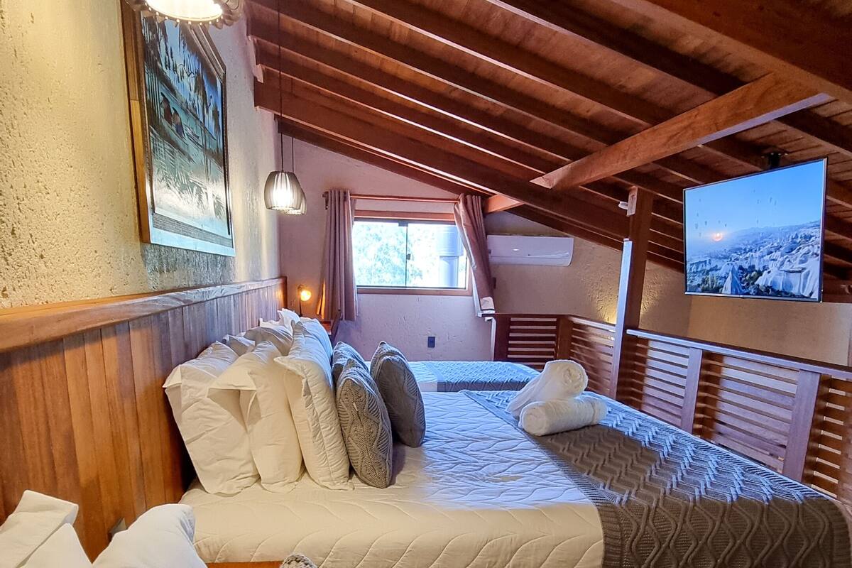 Ein gemütliches Zimmer mit Holzbalken, einem großen Bett mit vielen Kissen, einem Fernseher und einem Fenster mit Vorhängen.