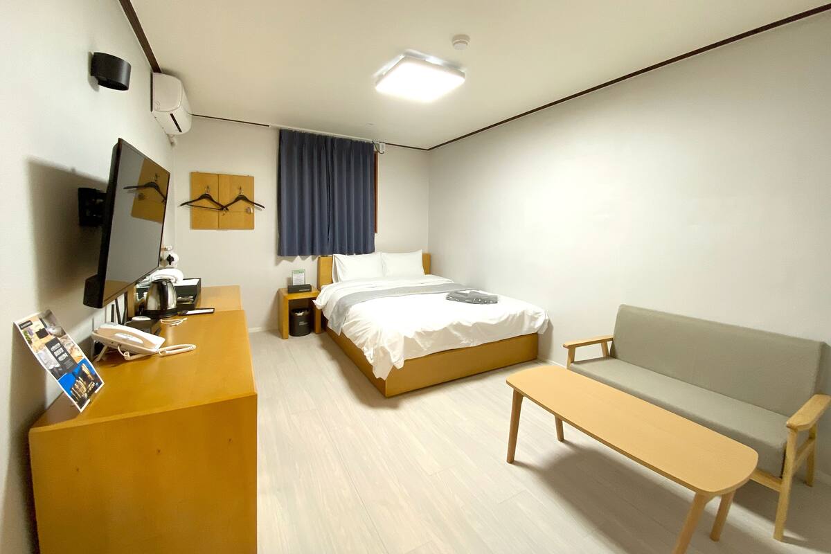 A hotel room with a bed, a desk with a telephone, a chair, a sofa, a small table, a mirror, a clock, and a window with curtains.