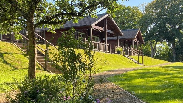 A wooden lodge with a porch, a staircase, and a well-maintained lawn. - Hollybush Lodges (Radstock)