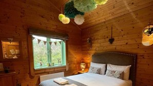 A wooden cabin bedroom with a large bed, a window with a view, and a hanging flower decoration. - Hollybush Lodges (Radstock)