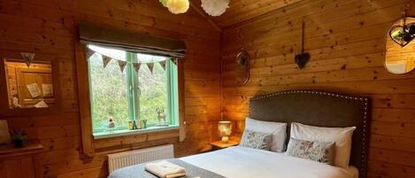 Quarto de cabana de madeira com uma cama grande, janela com vista e decoração floral pendente.