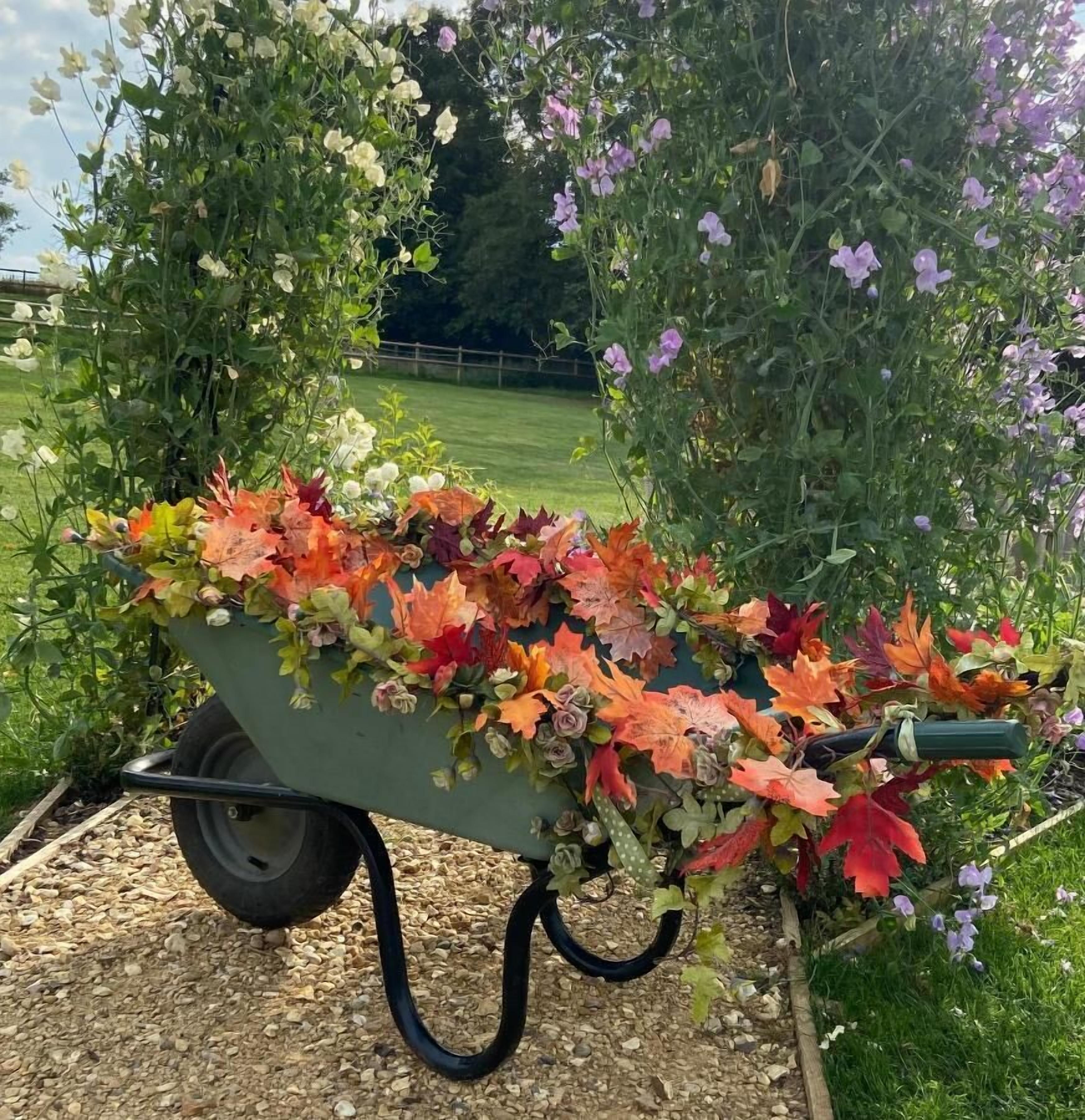Ein Schubkarren voller bunter Herbstblätter und Blumen, draußen auf einem Kiesweg mit einem grasbewachsen Feld und Bäumen im Hintergrund.