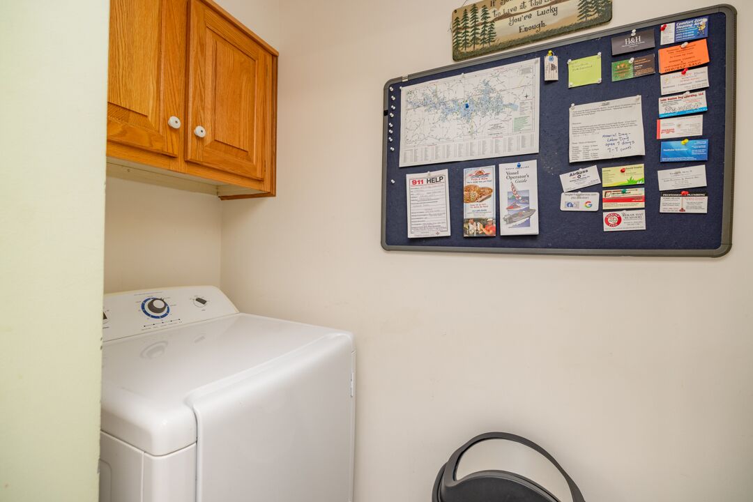 Een wasruimte met een witte wasmachine, een prikbord met diverse mededelingen en een houten kast erboven.