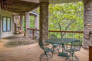 Una terraza cubierta al aire libre con un pilar de piedra, una mesa de metal con sillas, y una escalera de madera que sube a un nivel superior.