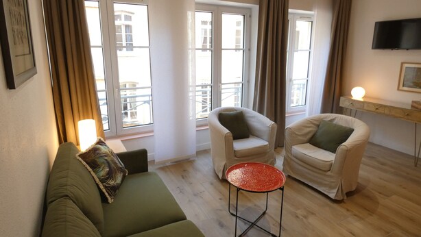 Een moderne woonkamer met een groene bank, twee witte fauteuils, een rode tafel en grote ramen met gordijnen.