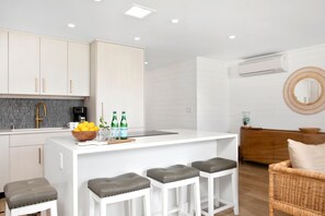 A modern kitchen with white cabinetry, a central island with stools, a coffee machine, and a bowl of fruit. - Maalaea Surf | Oceanfront 1 BR | Water Front | Car Incl. w/ 6+ Nights | MSR-F2 by KBM (Kihei)