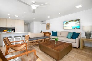Ein modernes Wohnzimmer mit einer Couch, einem Couchtisch und Sesseln, eine Küchenzeile mit einer Bar sowie ein Deckenventilator.