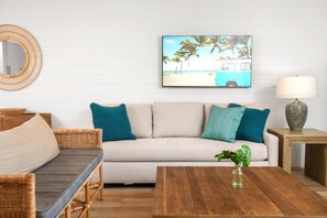 Un salon comprenant un canapé, une table basse, un fauteuil en osier, une lampe et un tableau représentant une scène de plage.