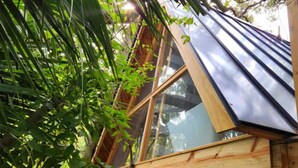 A wooden structure with a slanted roof and large windows, surrounded by greenery.