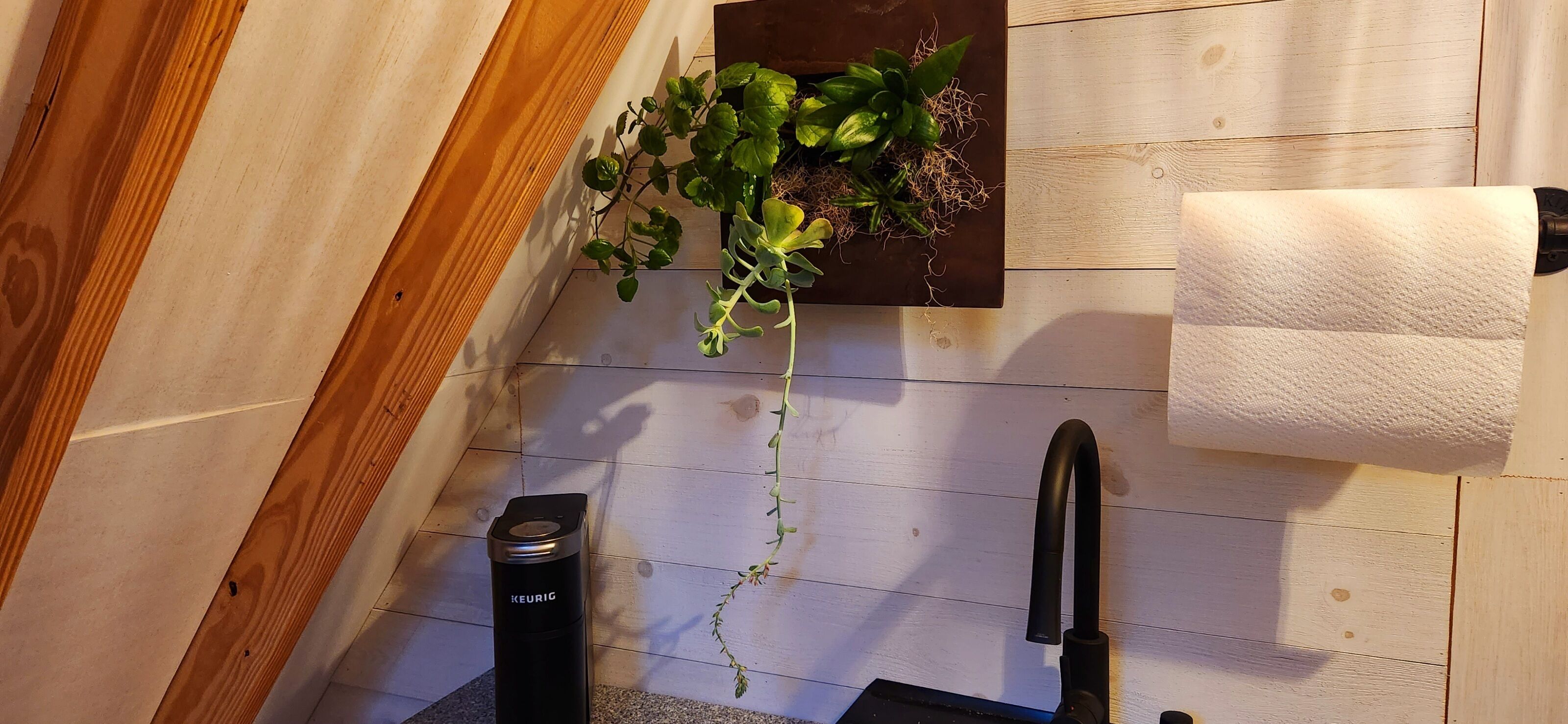 A corner of a room with a wooden floor, a black KEURIG coffee maker, a hanging plant with green leaves, and a white towel on a rack.