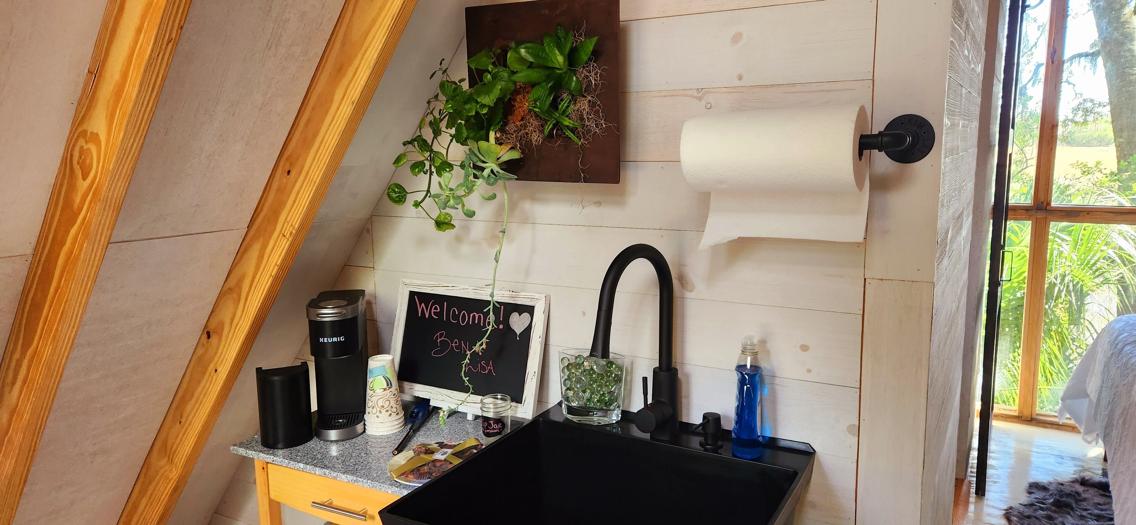 A kitchen with a black sink, a coffee maker, a chalkboard with a welcome message, and a window with a view of greenery.