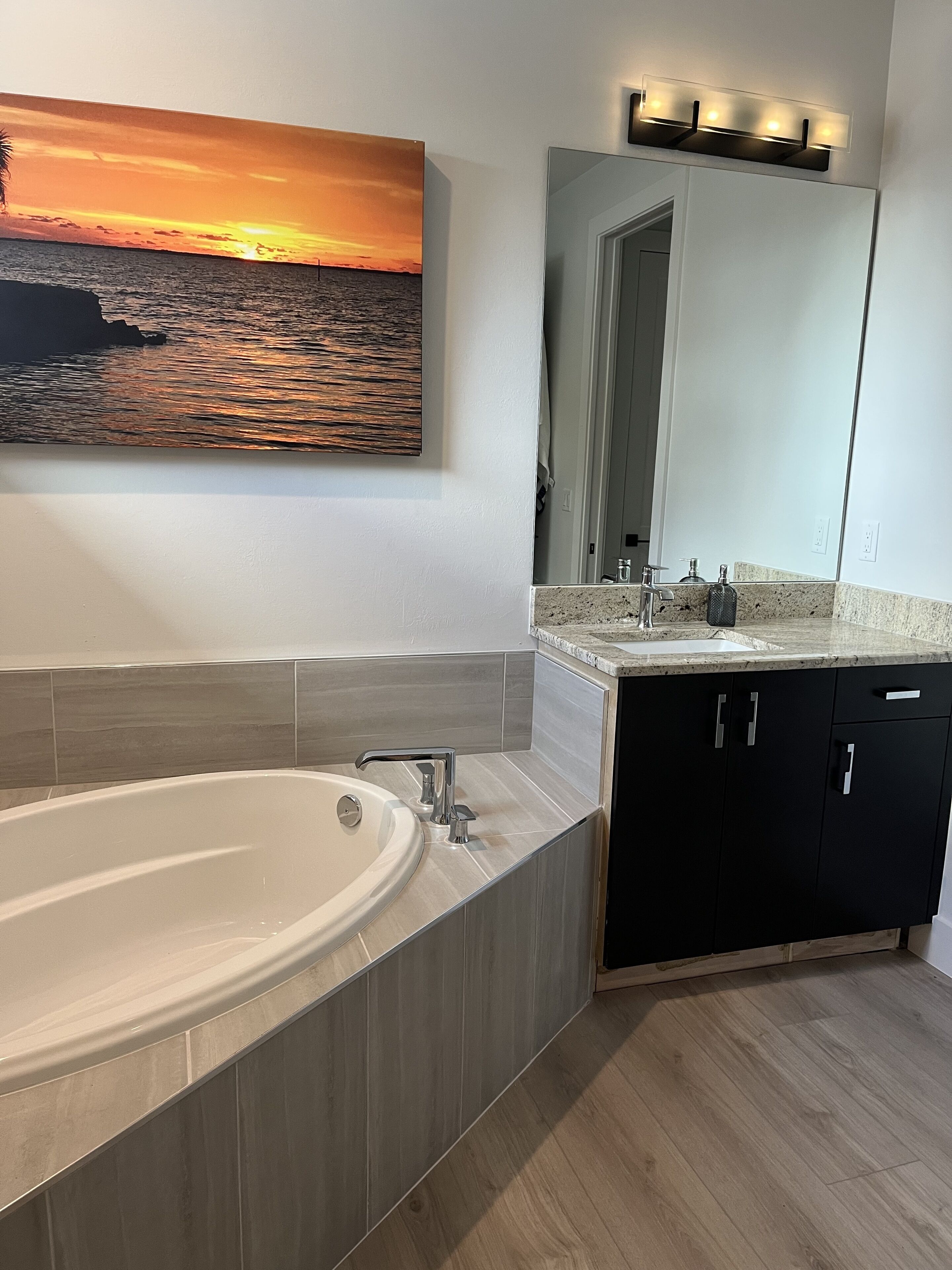 A modern bathroom with a large bathtub, a sink with a mirror above, and a painting of a sunset over the ocean on the wall.