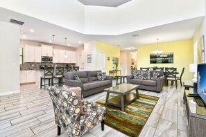 A spacious living area with a grey sofa, patterned armchair, and a coffee table. A dining area with a yellow wall and a chandelier is visible in the background.
