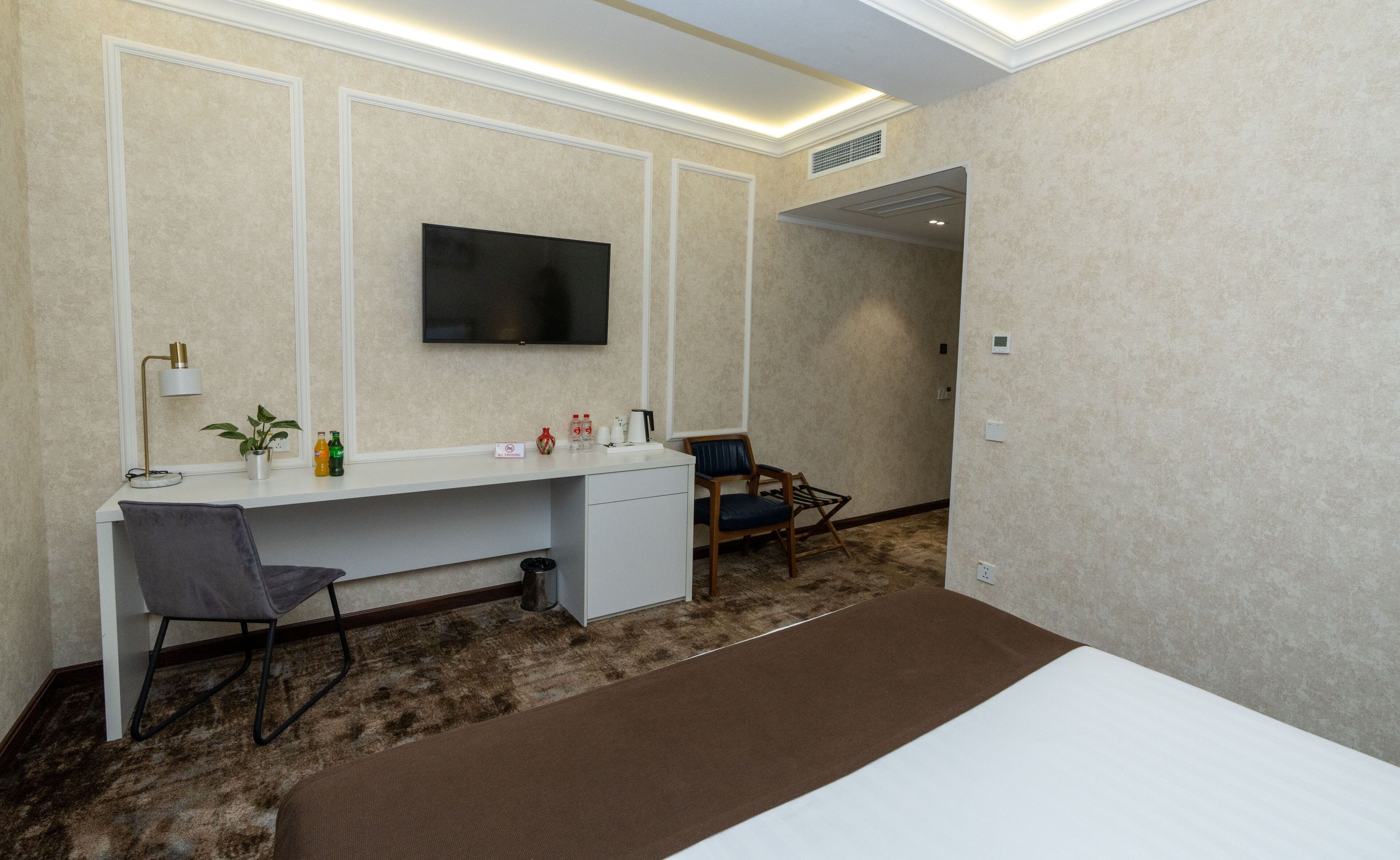 A hotel room with a bed, a desk with a chair, a TV mounted on the wall, and a small seating area with a chair and a table.