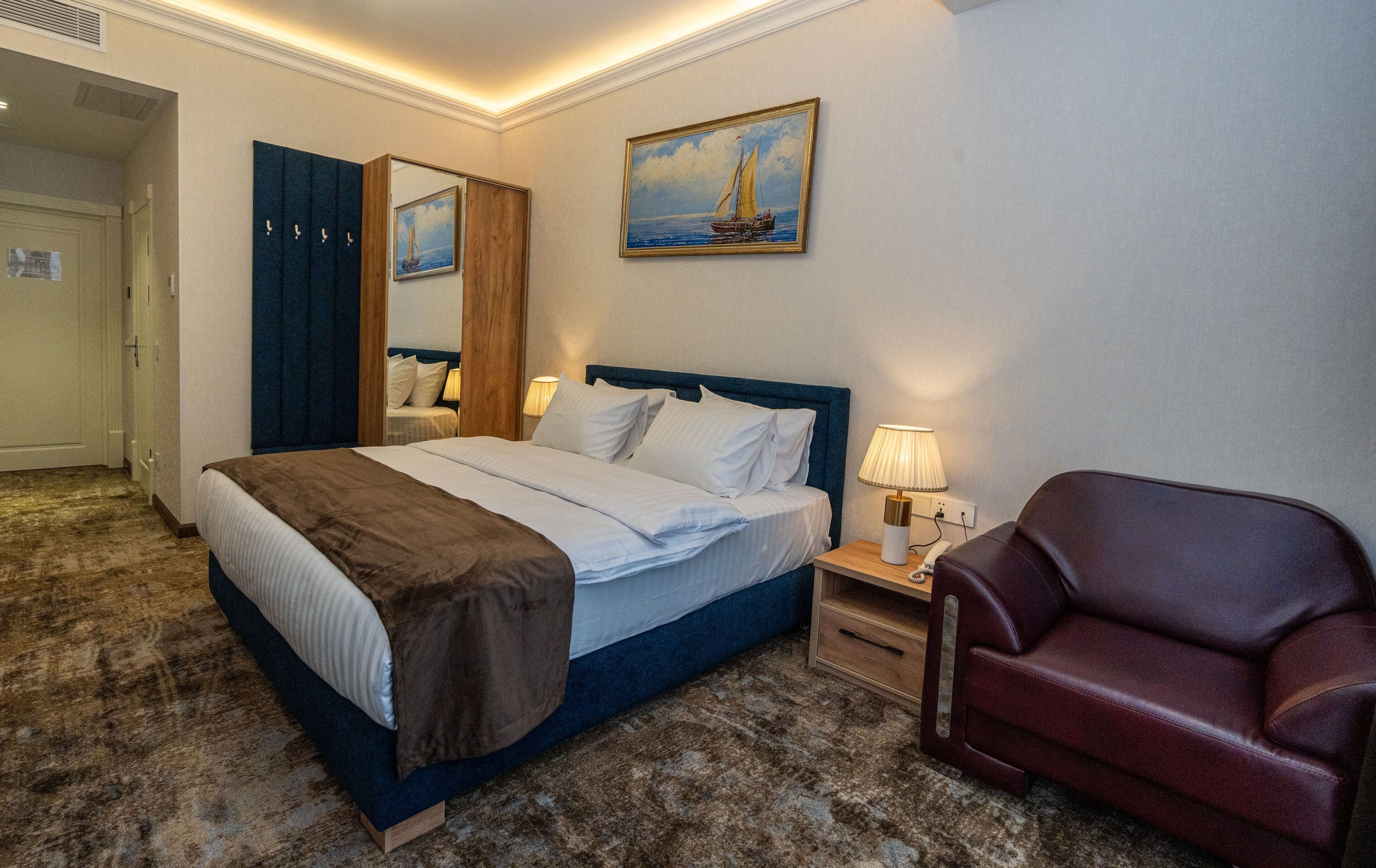Ein Hotelzimmer mit einem großen Bett, einem Nachttisch aus Holz, einem roten Sessel, einem Bild von einem Segelboot und einem Spiegel an der Wand.