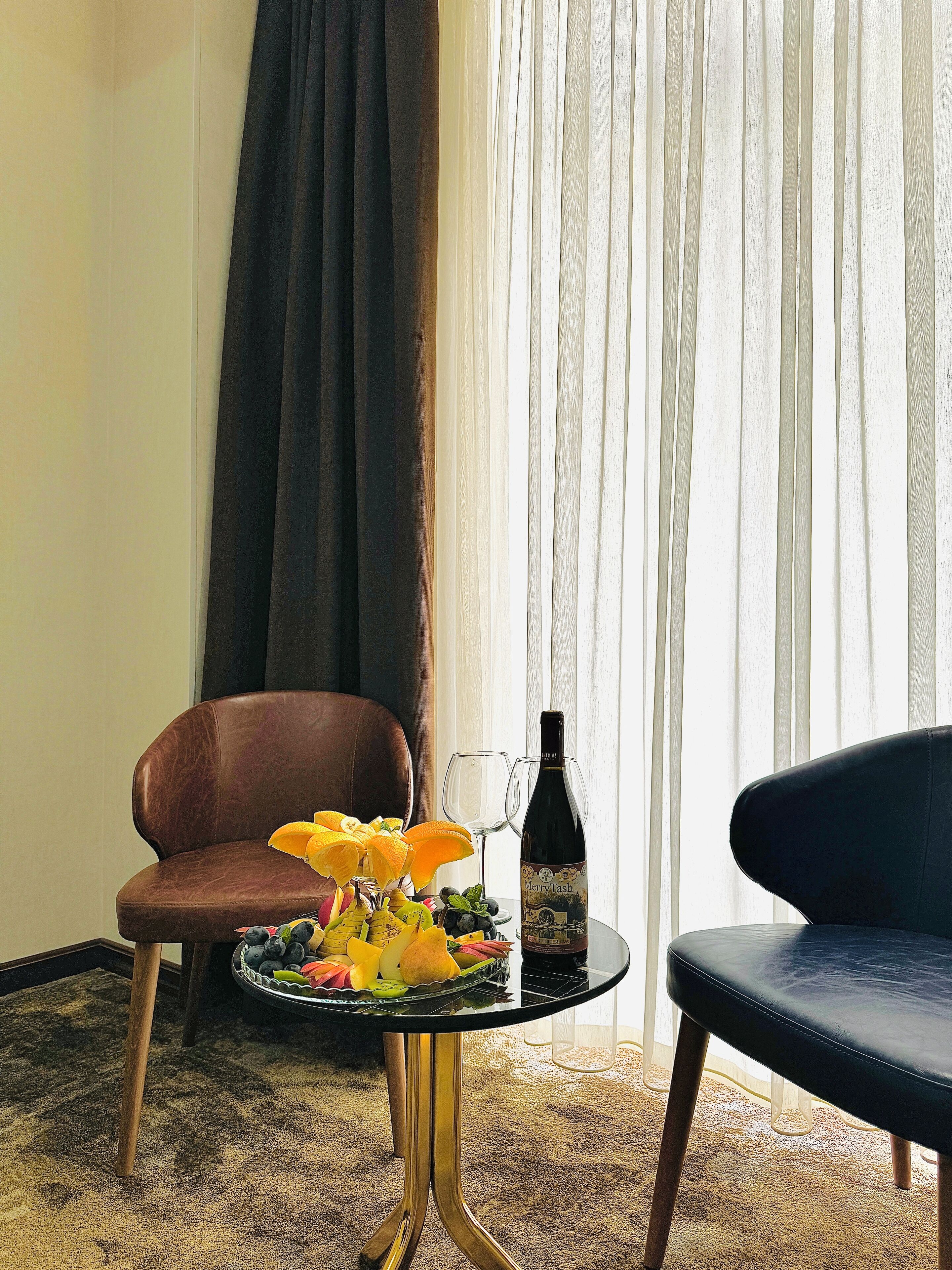A room with a brown leather chair, a black leather chair, a small table with a bottle of wine, glasses, and a fruit bowl.
