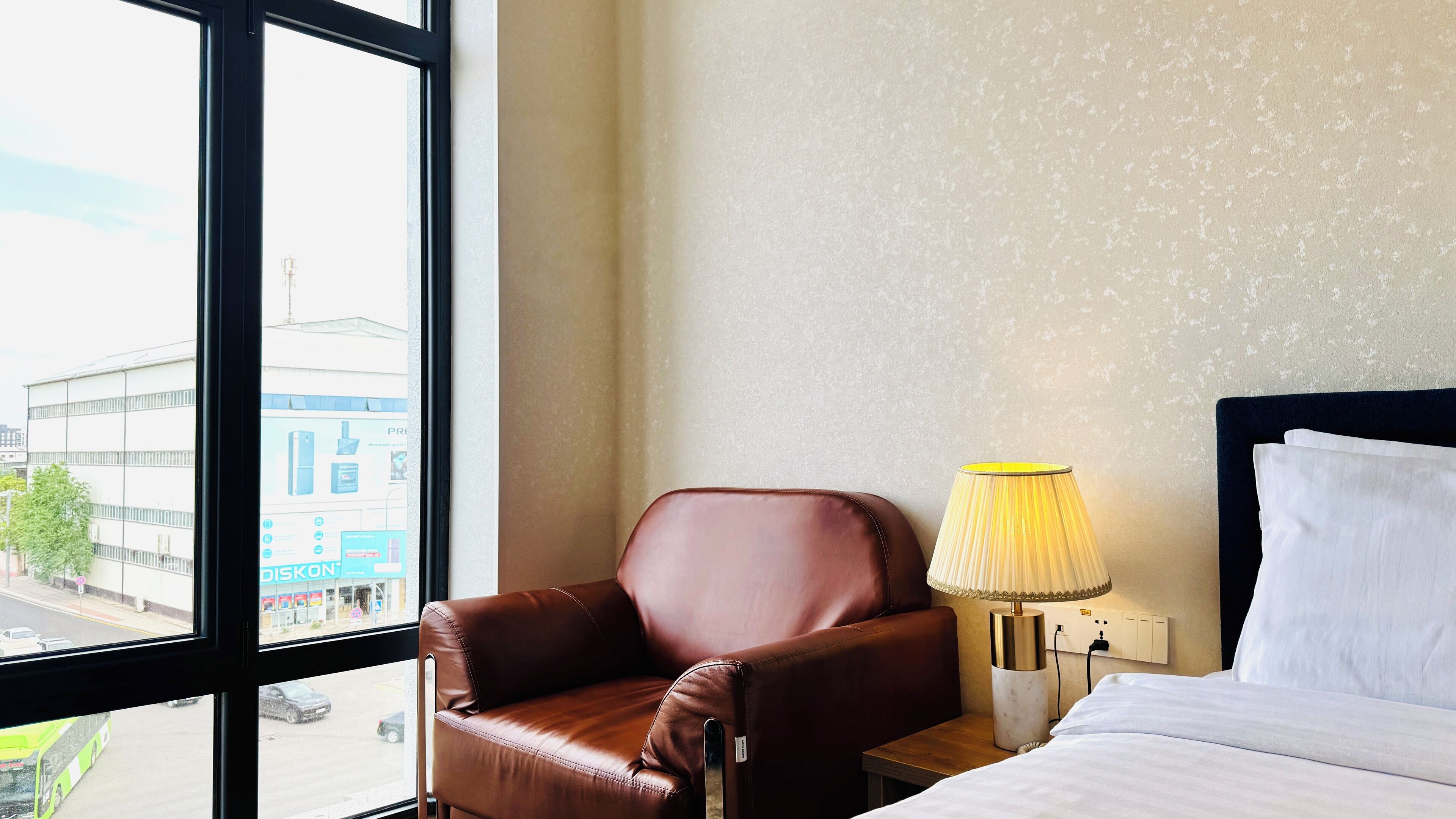 A hotel room with a leather armchair, a bedside lamp, a bed with white linens, and a large window offering a city view.