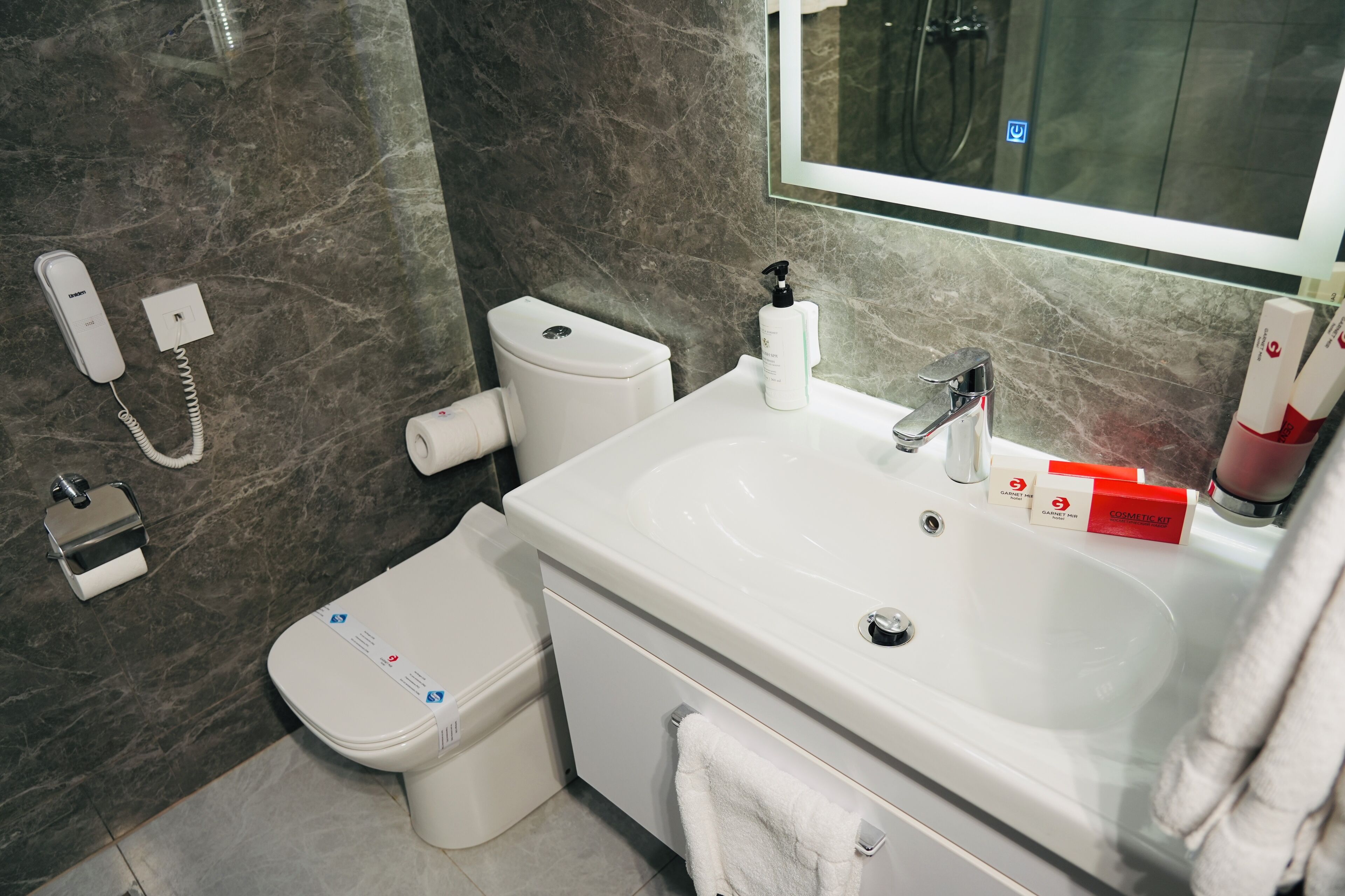Une salle de bain moderne avec un mur en marbre, un lavabo blanc, des toilettes et un téléphone.