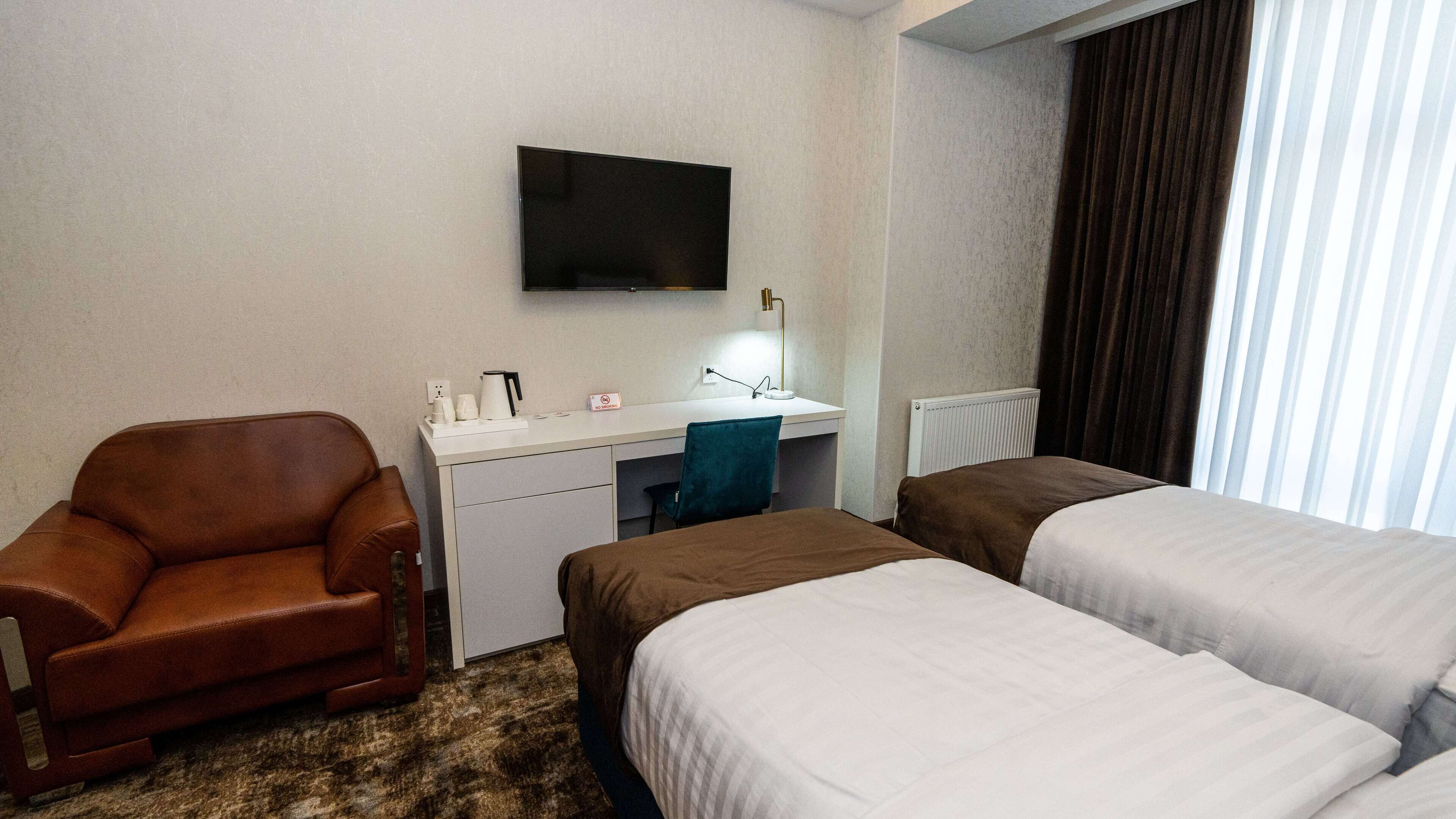 A hotel room with a brown leather chair, a desk with a flat-screen TV, a small sink, and two beds with white linens.